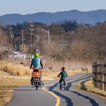 An adult and child on bikes moving away from the camera on a paved path with mountains and trees in the background.