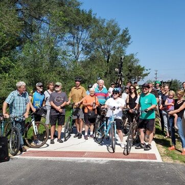 A large group of people of all ages, some on bikes and some on foot posing for the photo at the beginning of a paved rural multiuse path.