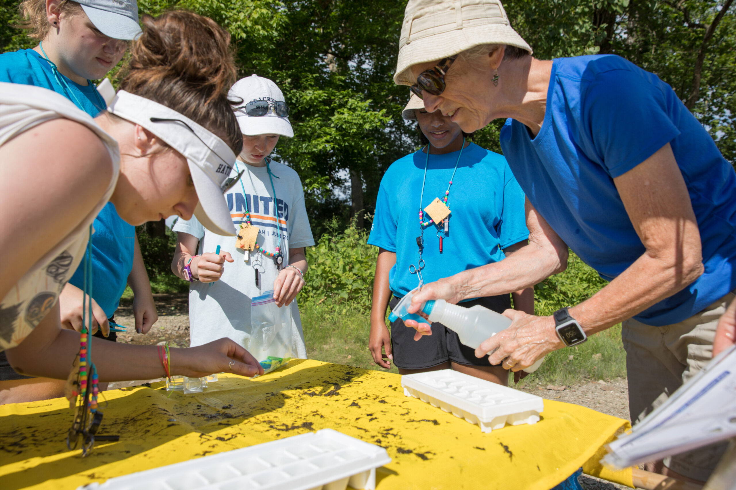 A group of women and young girls lean over a table of stream sediment apparently sorting things into ice cube trays for analysis.