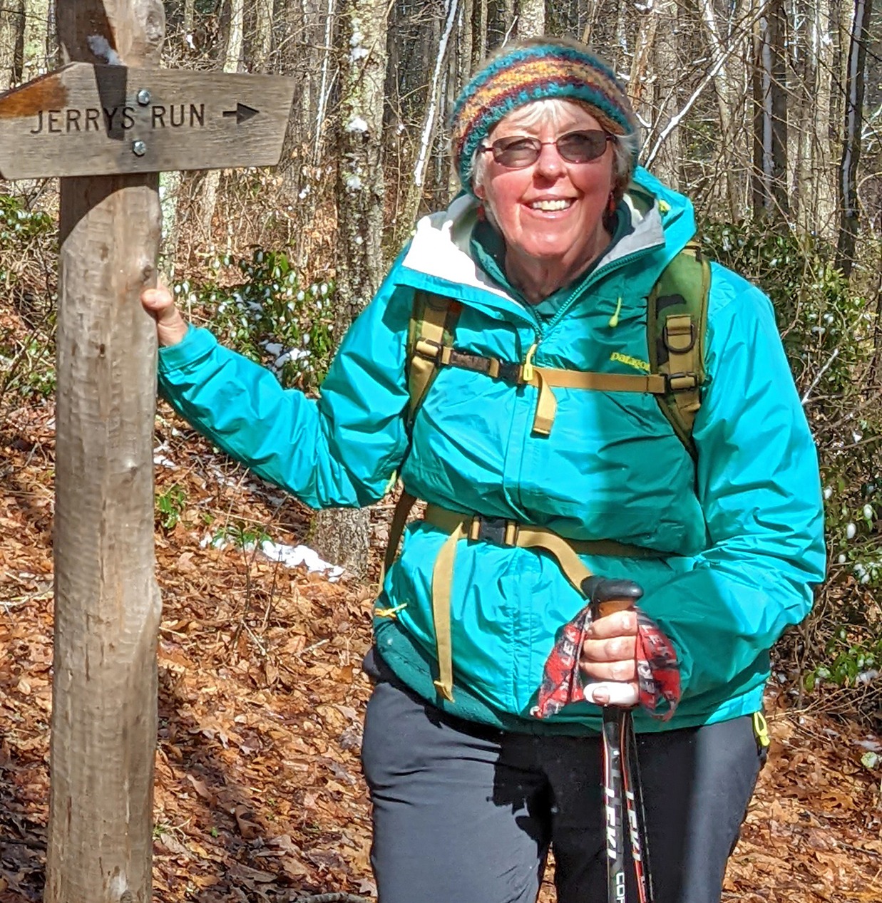 Lynn Cameron wearing cold weather hiking gear and holding hiking polls leaning on a sign post that says 'Jerry's Run.'