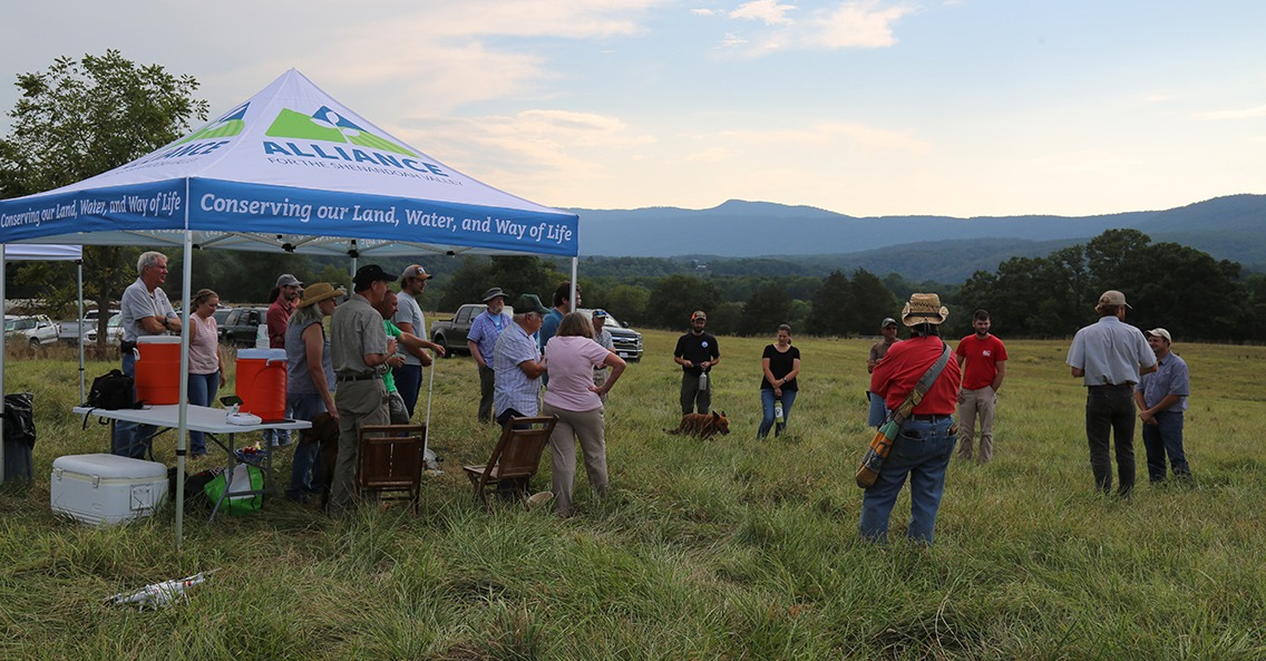About 25 people gather around an Alliance for the Shenandoah Valley popup tent in a field. The tent says 'conserving our land, water, and way of life.'
