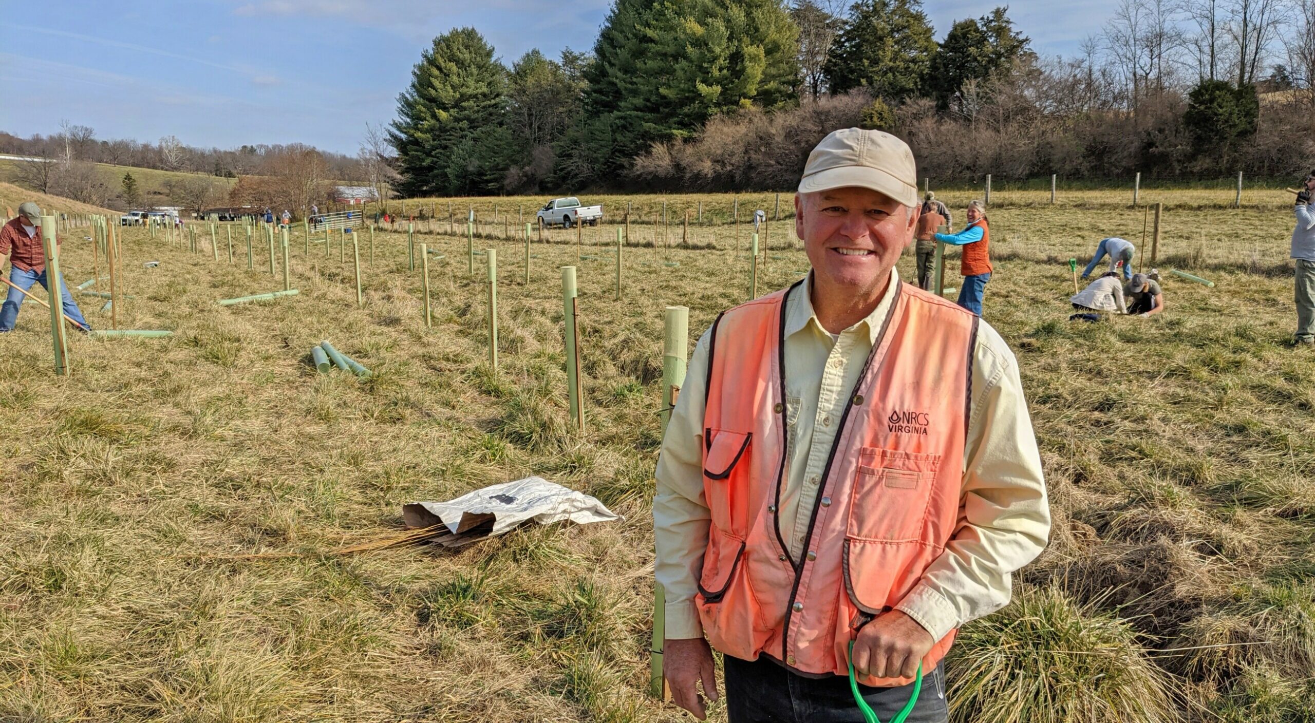Bobby Whitescarver standing in his field with volunteers planting seedlings in the background.