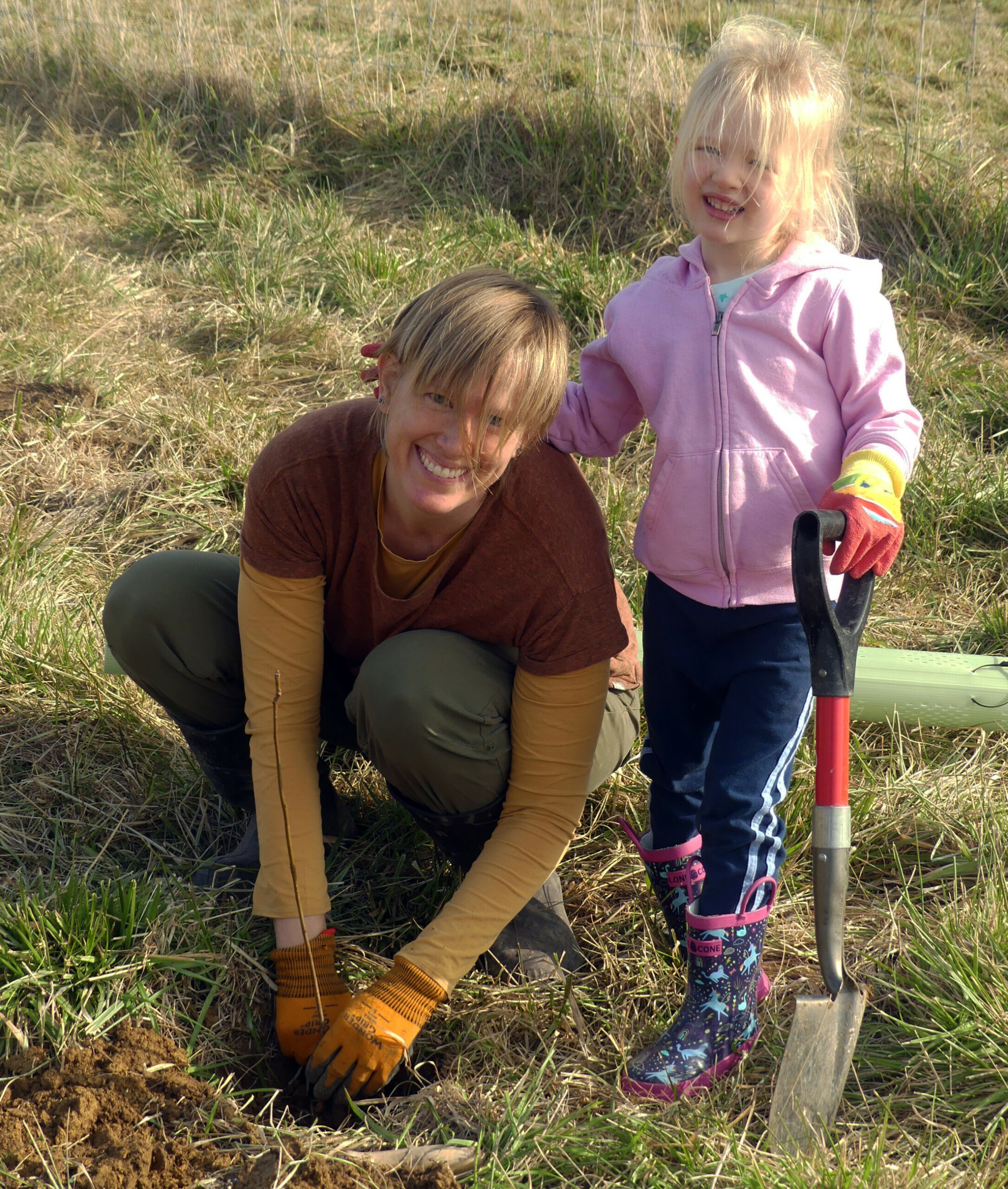 A mother and daughter smile as the mother plants a sapling and the daughter holds a small shovel.