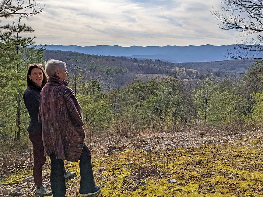 Two women stand on an overlook covered with lime green moss with a view of green trees and fields fading to blue mountains and a hazy white sky.