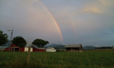 Double rainbow in a hazy sky over a lush green farm field with red barns in the distance.