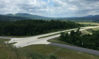 A massive 4-lane divided highway intersects with a rural unmarked road in mountainous countryside.