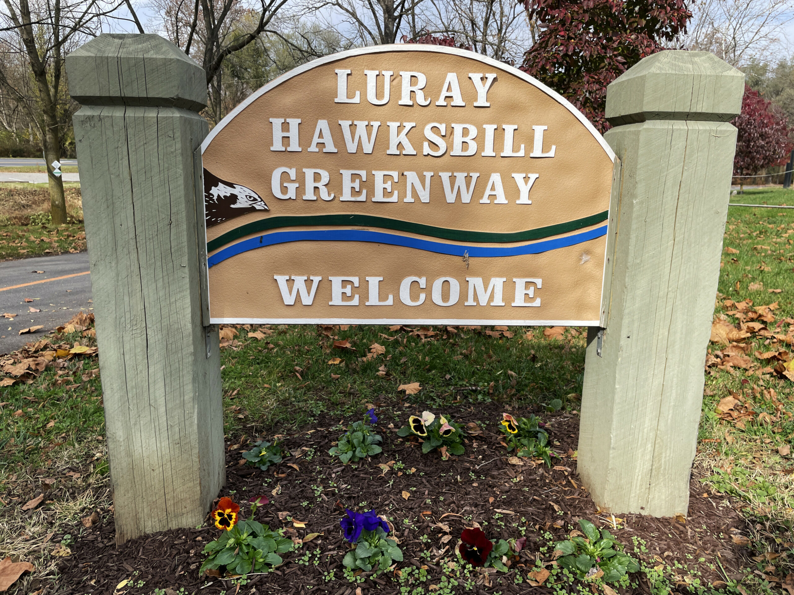 Wooden greenway welcome sign between two concrete posts with pansies planted underneath and a paved path in the background.