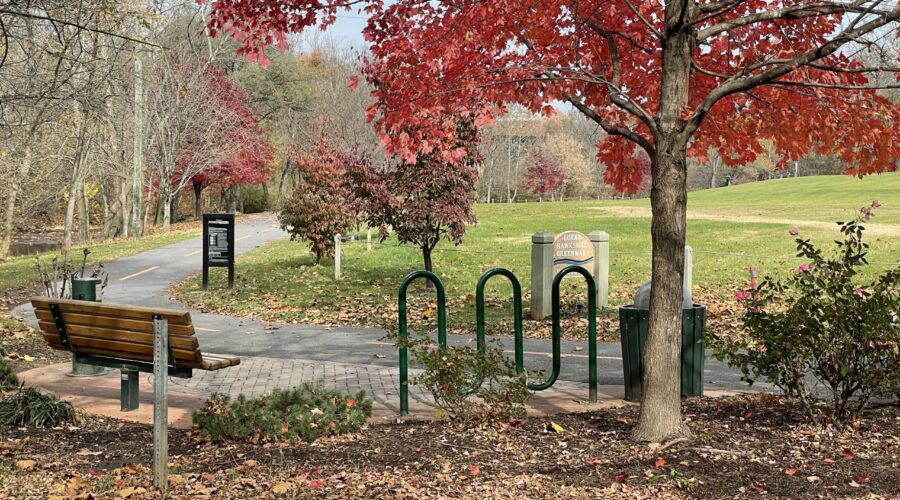 A paved walking path winds around a bend with a bench and bike racks under a tree with red fall leaves.