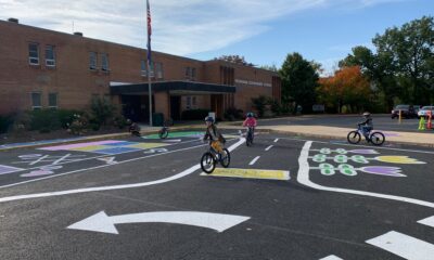 Children on bikes ride on pavement that has been painted to resemble a roadway in front of a school.