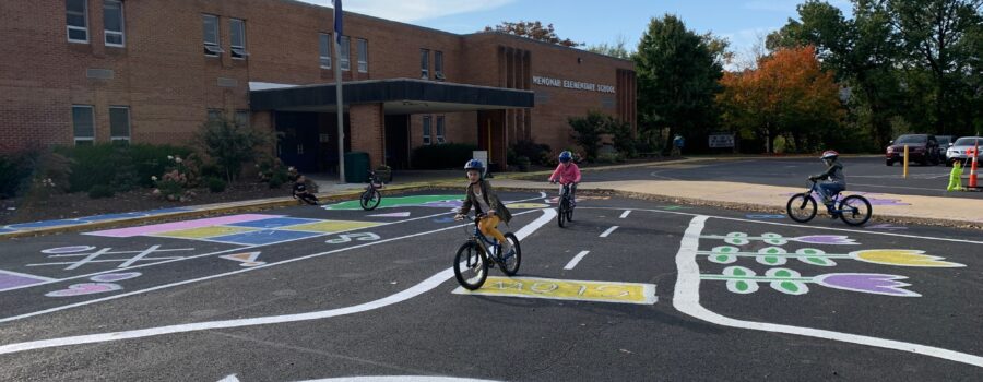 Children on bikes ride on pavement that has been painted to resemble a roadway in front of a school.