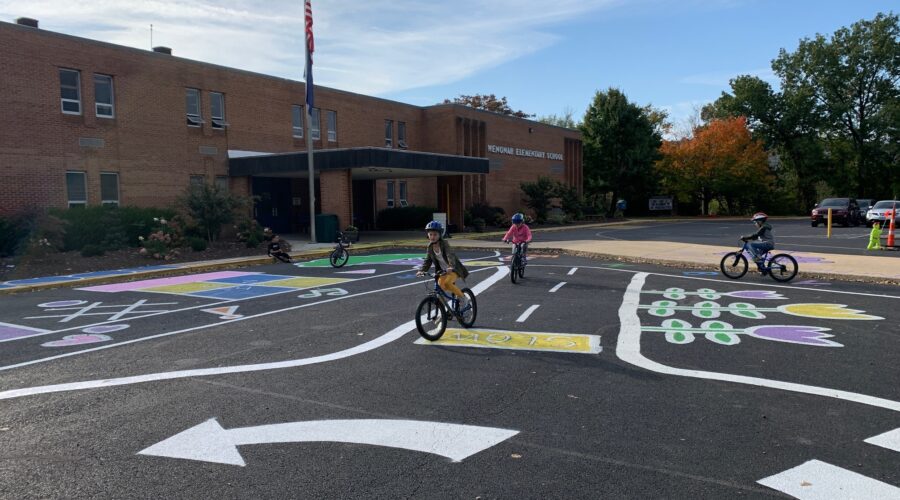 Children on bikes ride on pavement that has been painted to resemble a roadway in front of a school.