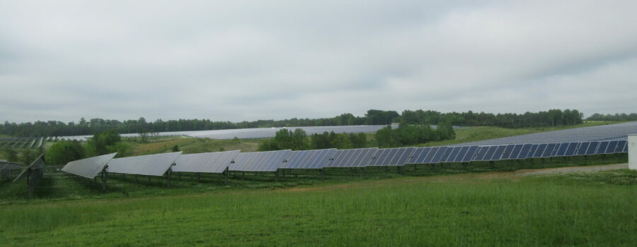 A solar array tucked away between green shrubs and fields under a cloudy sky.