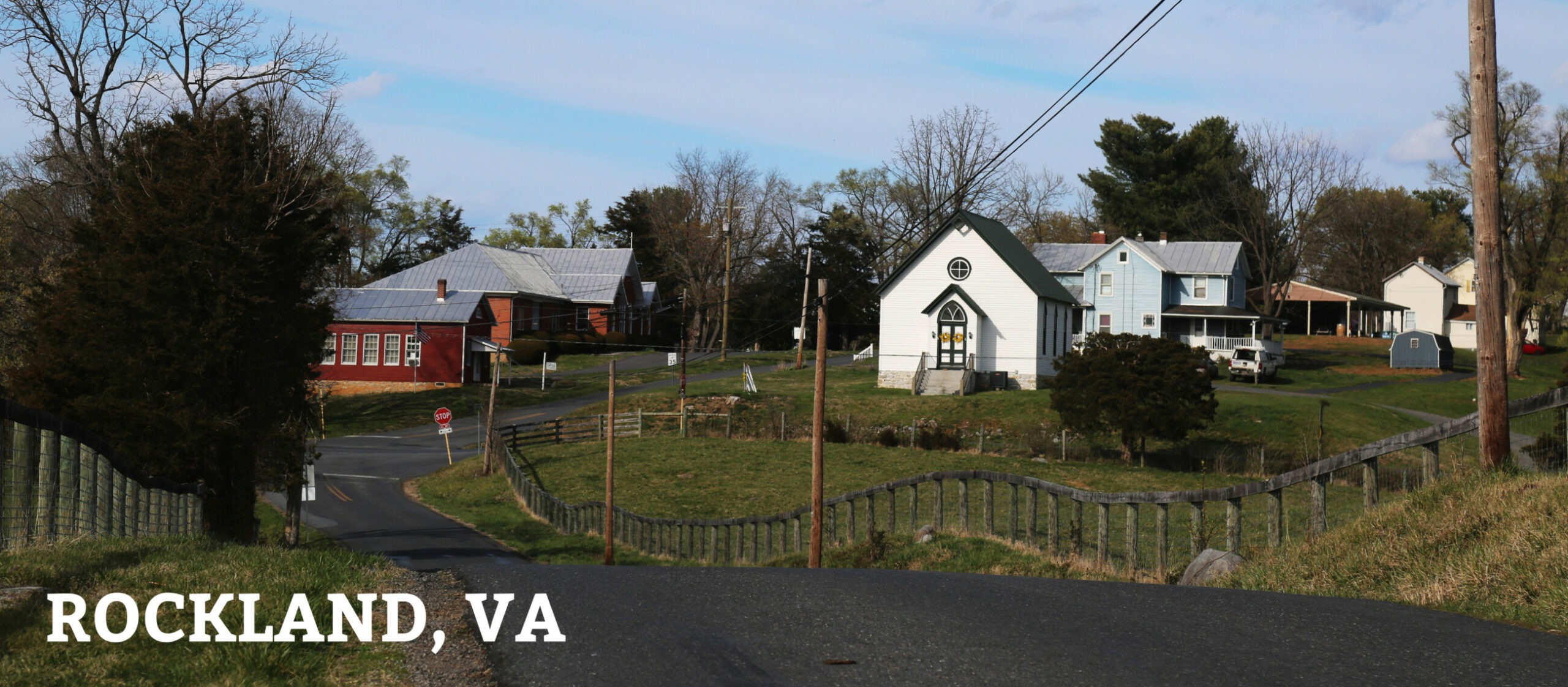 Historic churches and homes dot a narrow rural roadside bordered on both sides by a wooden farm fence. 