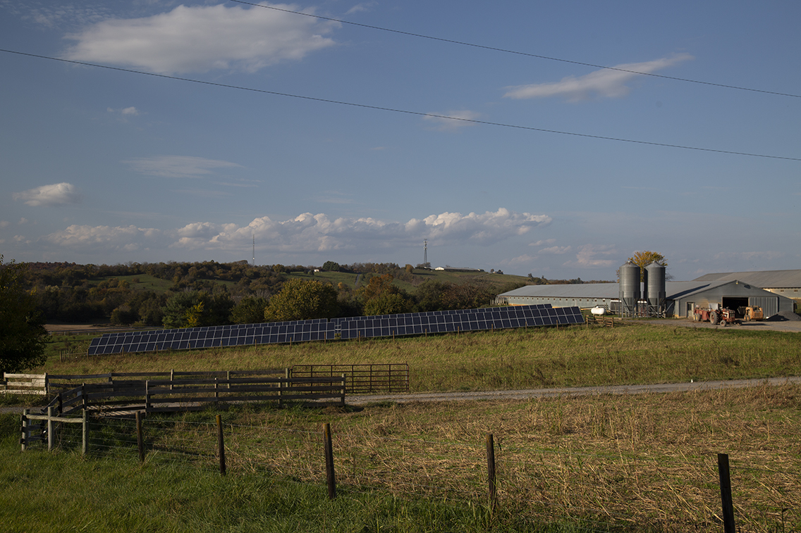 A barn and silos neighboring a small solar array with fall farm fields in the foreground, rolling tree-covered hills in the background all under a blue sky dappled with clouds.