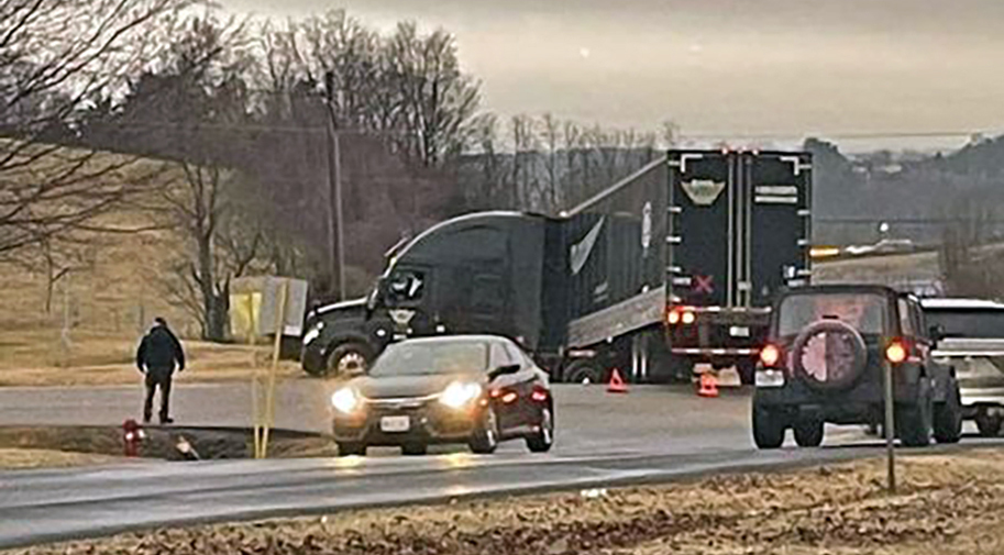 A large black 18-wheel semi truck blocks traffic on a two-way divided highway on a hazy, grey winter day.