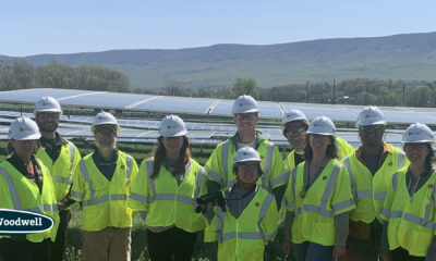Ten people pose in front of a solar array in a farm field wearing bright yellow safety vests and white hard hats.