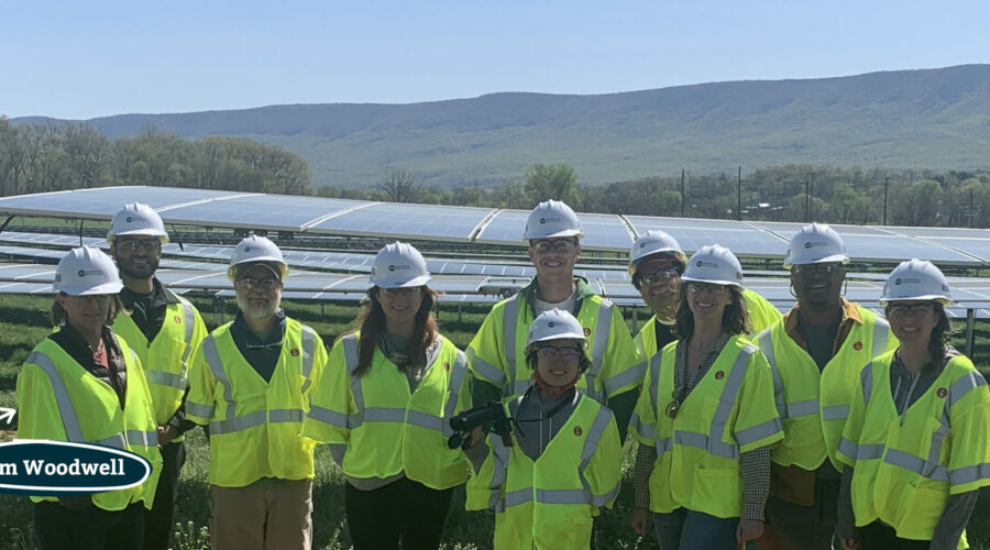 Ten people pose in front of a solar array in a farm field wearing bright yellow safety vests and white hard hats.