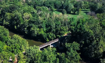 An areal view of a trestle crossing a wide river surrounded by green leafy trees.