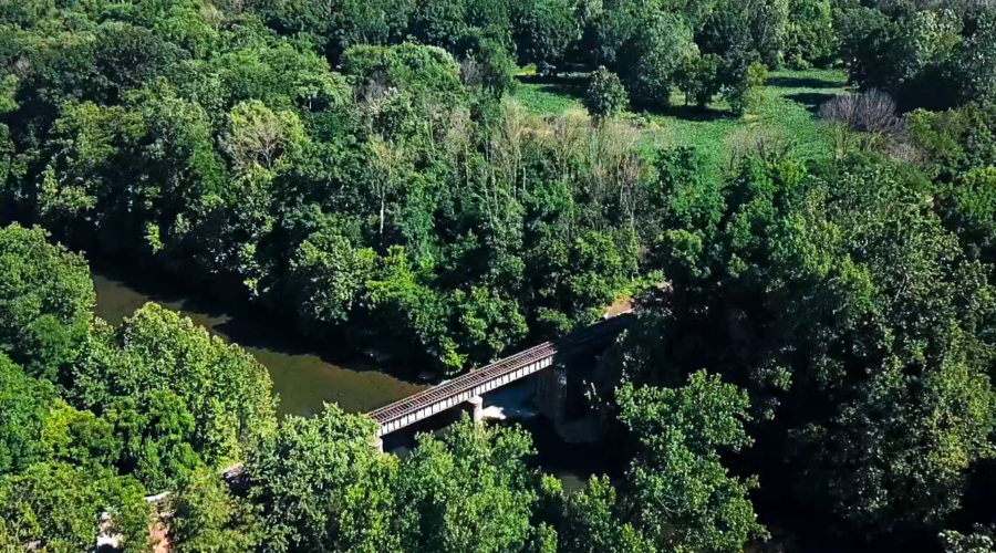 An areal view of a trestle crossing a wide river surrounded by green leafy trees.