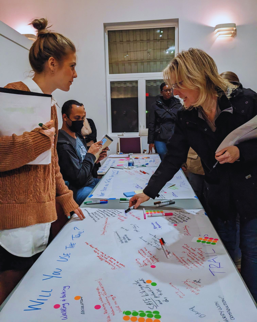 Two women lean over a table covered in paper written with comments in multi-colored marker and dots.