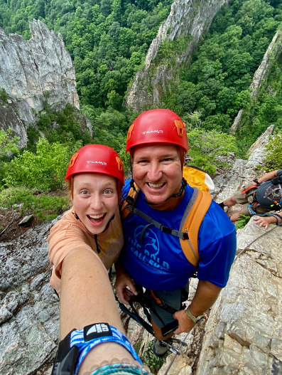 A man and woman wearing bright red helmets and climbing gear pose for a selfie on some steep looking rocks.