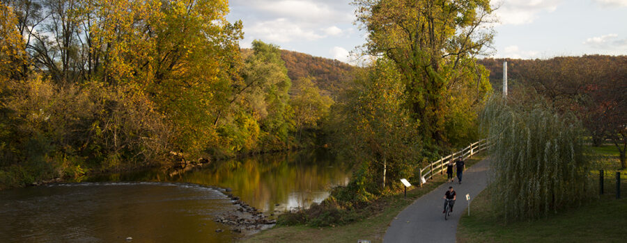 A fall scene of the South River running alongside the paved South River Greenway with cyclist passing a couple walking toward the camera.