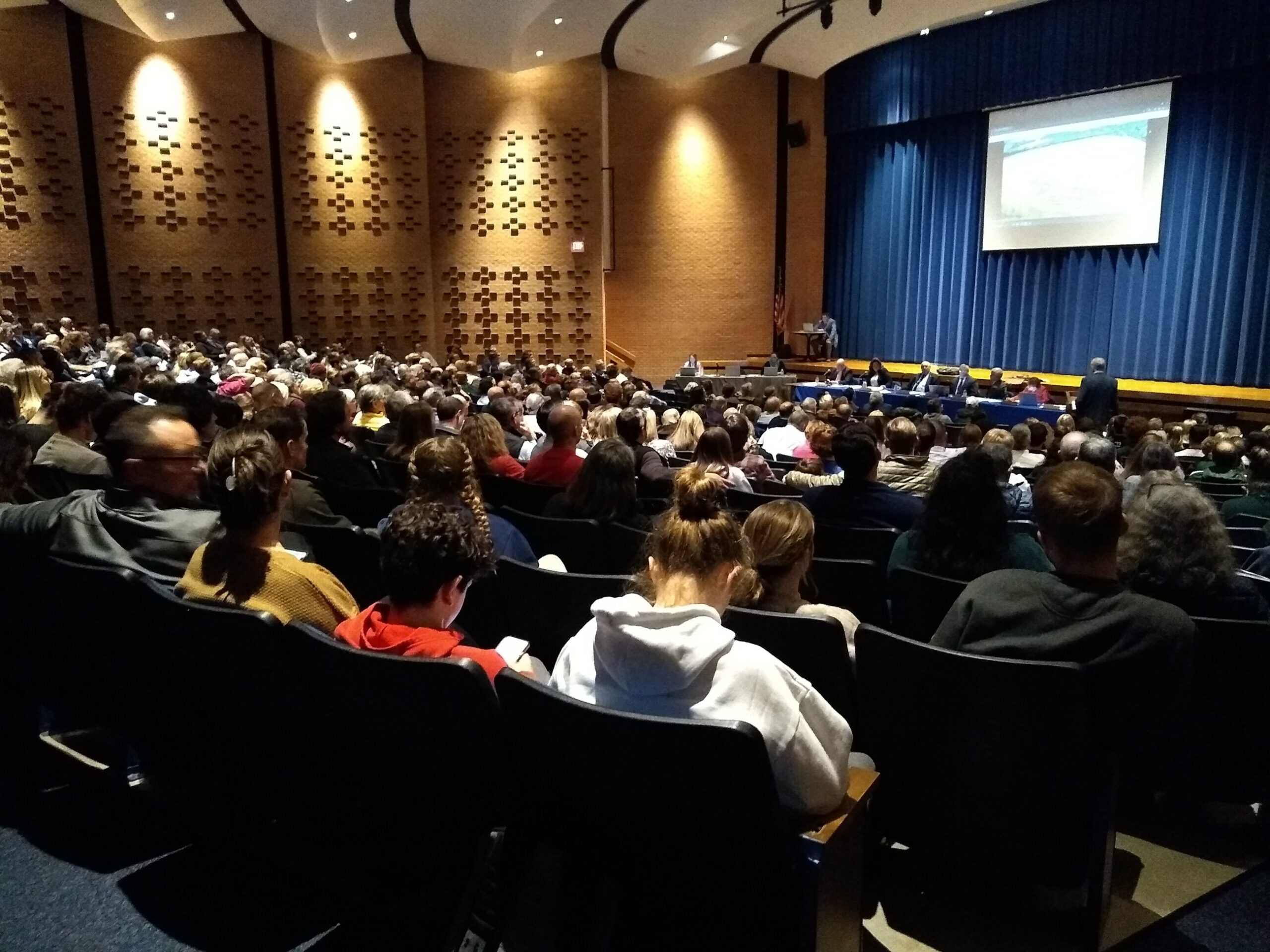 A wide shot of an auditorium filled with seated people with a governing body in front and a screen presentation against a closed blue curtain.
