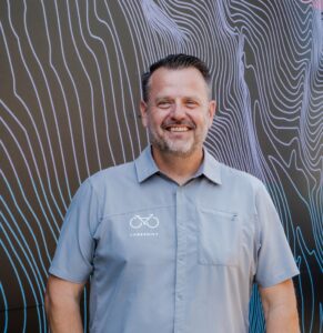 Laneshift CEO Ryan Hale poses with a smile for a camera. He's wearing a light blue shirt with a bicycle icon a standing in front of a topographically decorated brown wall.
