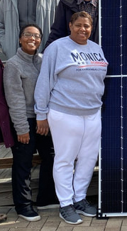 Two Black women pose smiling at the camera beside a solar panel.