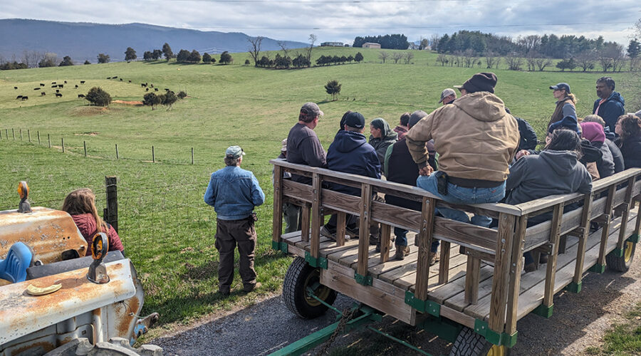 People sit in a wooden wagon behind a tractor looking off into the distance over a green field under a cloudy sky.